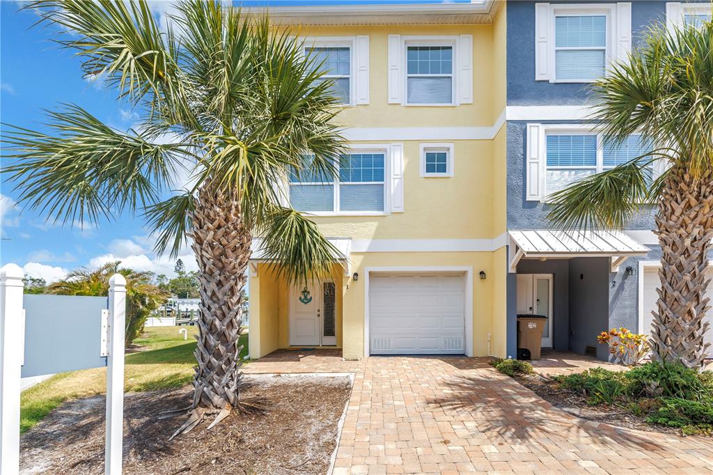 A three-story yellow townhouse with a garage, brick paver driveway, and palm trees in front — 100 WINDRUSH BOULEVARD UNIT 1, INDIAN ROCKS BEACH, FL