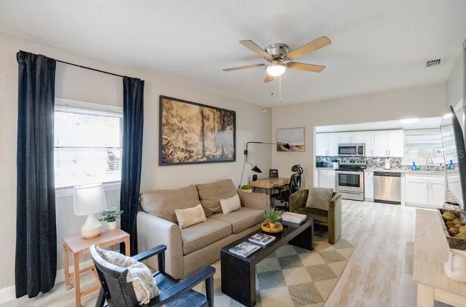 A living room with a sofa, coffee table, and ceiling fan opens into a white kitchen — 8311 N NEWPORT AVENUE, TAMPA, FL