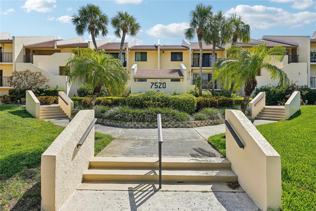 A multi-story apartment building with a central entrance, stairs, and palm trees in front — 7520 SUNSHINE SKYWAY LANE S UNIT T18, ST PETERSBURG, FL