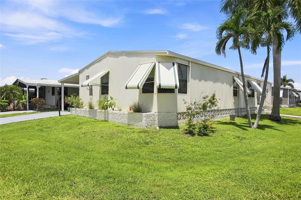 A light-colored mobile home with metal awnings sits on a grassy lot with palm trees — 252 LOON LANE UNIT 252, NAPLES, FL