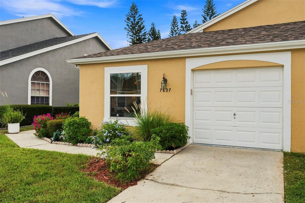A yellow house exterior features a single-car garage, a window, and a small front garden — 7637 BALHARBOUR DRIVE, NEW PORT RICHEY, FL