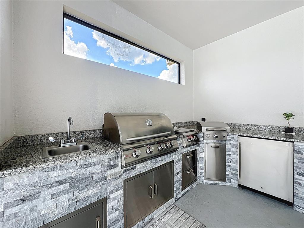 An outdoor kitchen features a stainless steel grill, sink, refrigerator, and stone-tiled cabinetry — 15084 CUDDY COURT, ORLANDO, FL