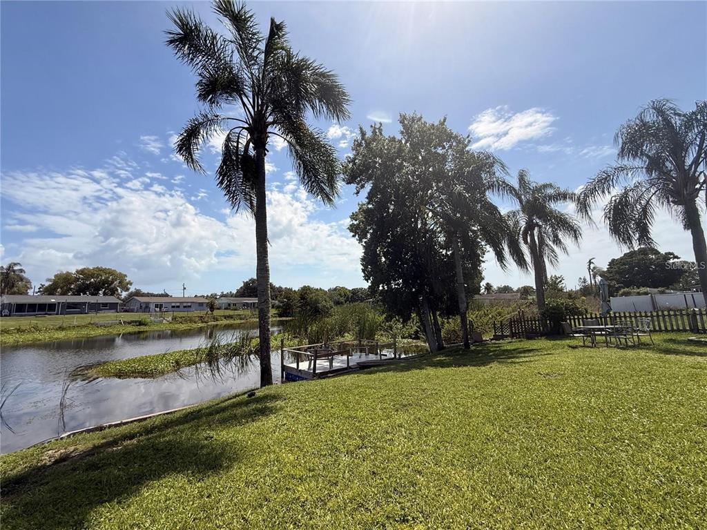 A grassy backyard features a small wooden dock extending into a pond with palm trees — 5144 CHIME WAY, HOLIDAY, FL
