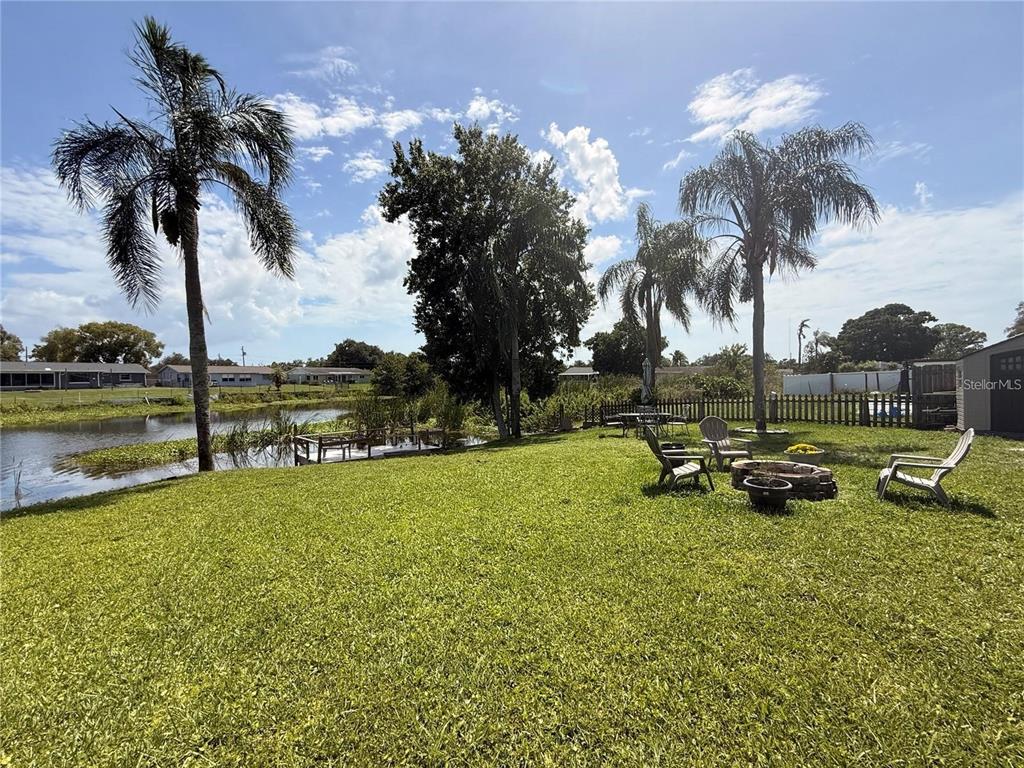 A grassy backyard features a small pond, palm trees, a fire pit, and patio furniture — 5144 CHIME WAY, HOLIDAY, FL