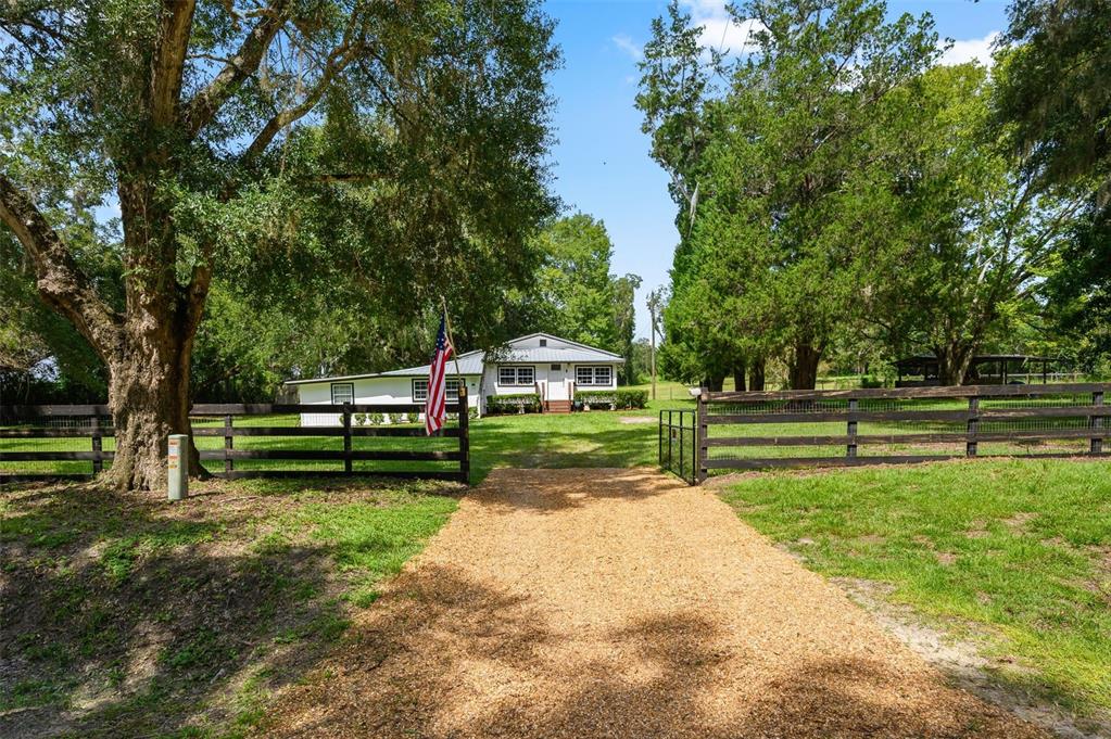 A gravel driveway leads to a white house behind a black wooden fence and trees — 6218 SW 202ND STREET, NEWBERRY, FL
