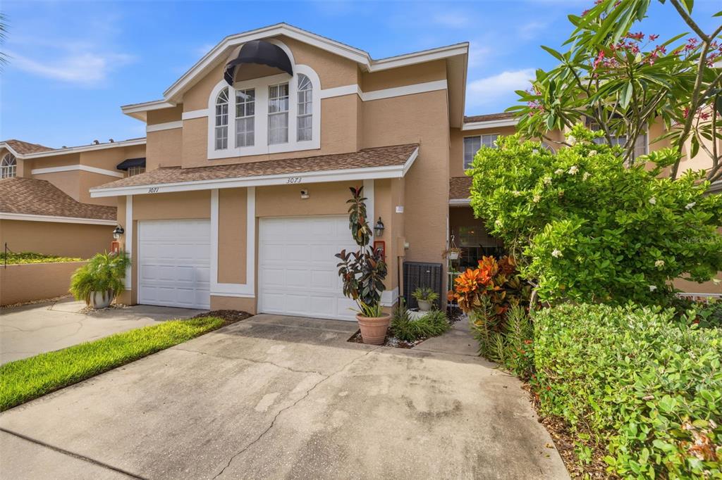 A two-story townhouse with a tan stucco exterior, two garage doors, and lush landscaping — 3073 BRANCH DRIVE, CLEARWATER, FL