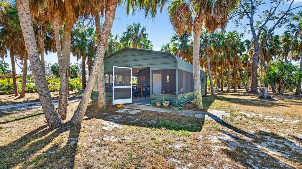 A green metal shed with a screened-in porch sits among palm trees on sandy ground — 2619 RIVER BEND DRIVE, RUSKIN, FL