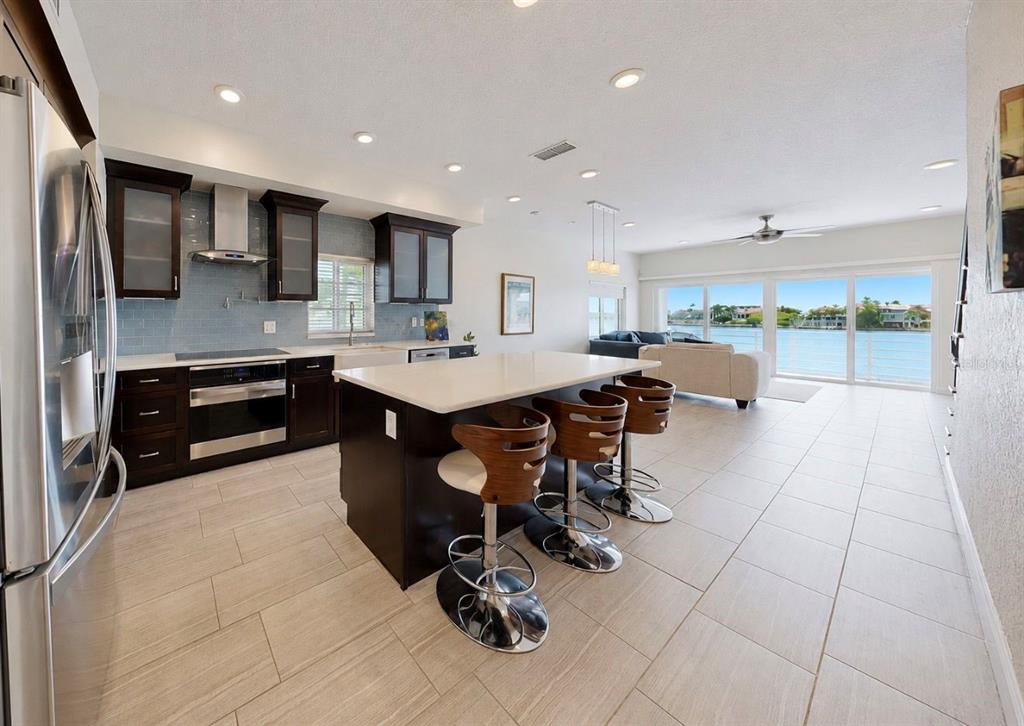 A modern kitchen with dark cabinets and a central island overlooks a living area and water — 4639 MIRABELLA COURT, ST PETE BEACH, FL