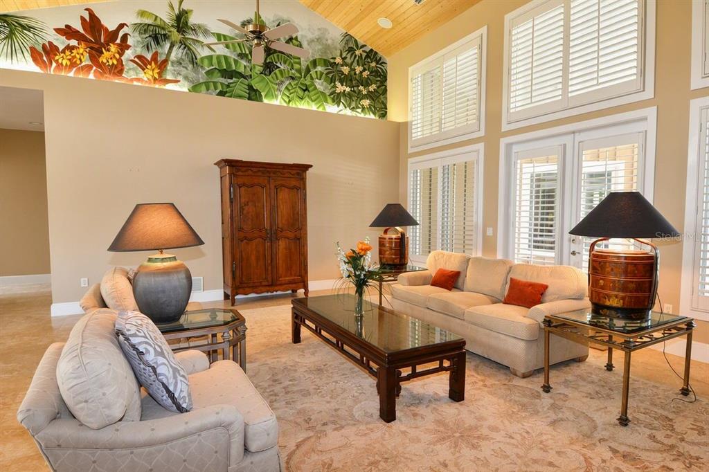 A living room with a tropical mural, high ceilings, beige furniture, and glass-topped tables — 630 N RIVER ROAD, VENICE, FL