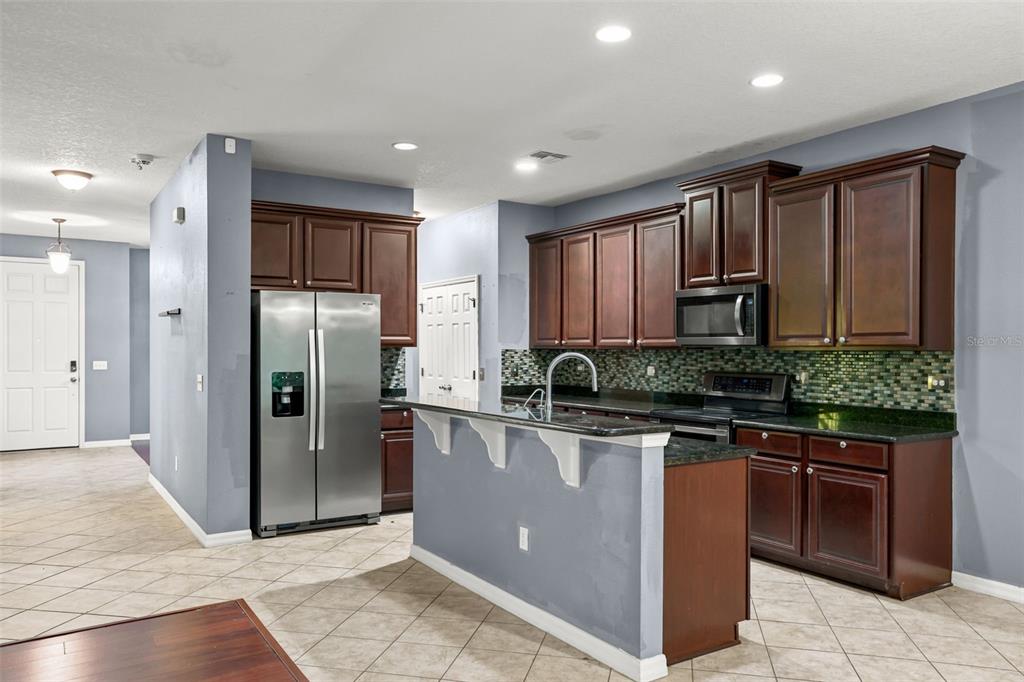 A kitchen features dark wood cabinets, granite countertops, a stainless steel refrigerator, and tile flooring — 925 COUNTS CREST CIRCLE, APOPKA, FL