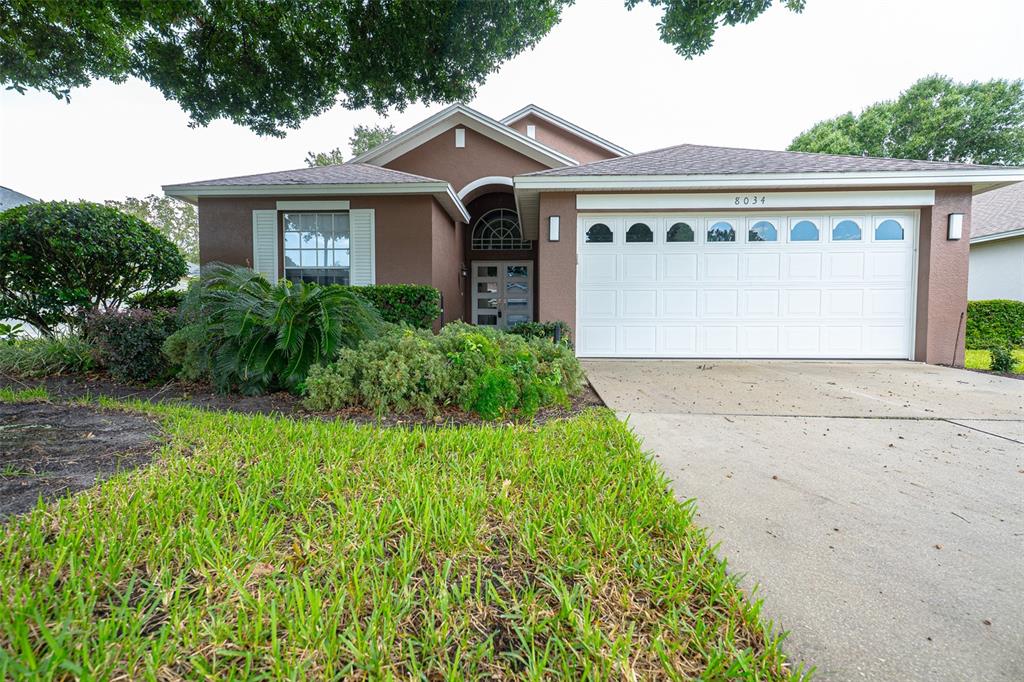 A brown single-story house features a white garage door, a front entrance, and landscaped yard — 8034 ARCADIAN COURT, MOUNT DORA, FL