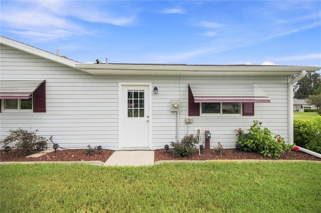 A light gray house exterior features a white door, two windows with awnings, and landscaping — 17790 SE 101ST COURT, SUMMERFIELD, FL
