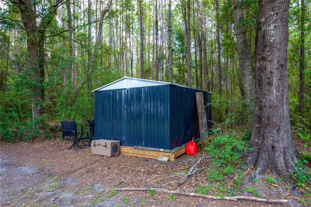 A dark blue metal storage shed sits on a wooden platform in a wooded area — 13328 NE 230TH COURT, RAIFORD, FL