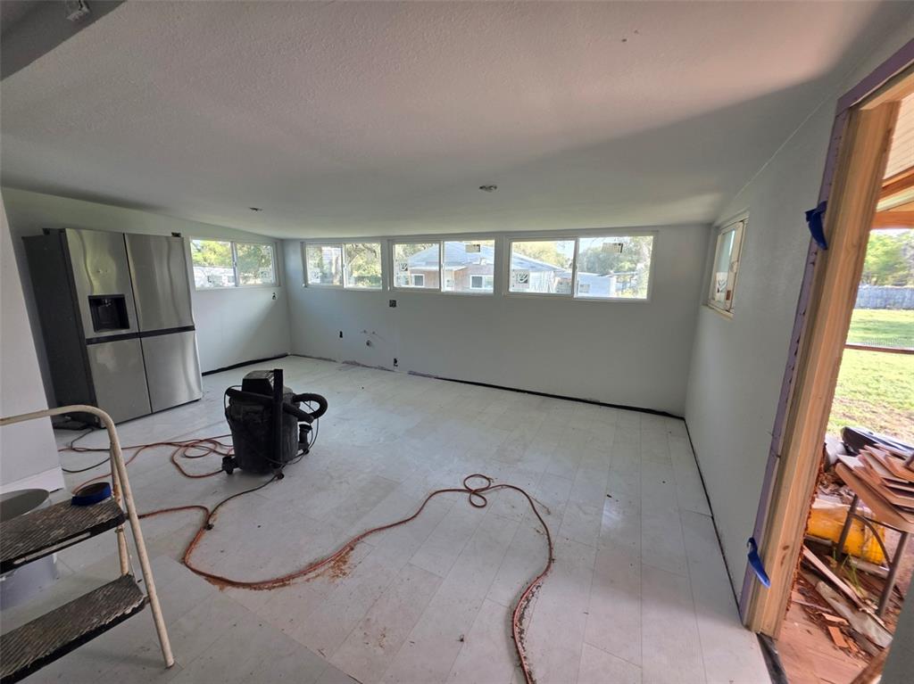 An unfinished room features light-colored flooring, white walls, multiple windows, and a stainless steel refrigerator — 37414 NORTHSIDE DRIVE, ZEPHYRHILLS, FL