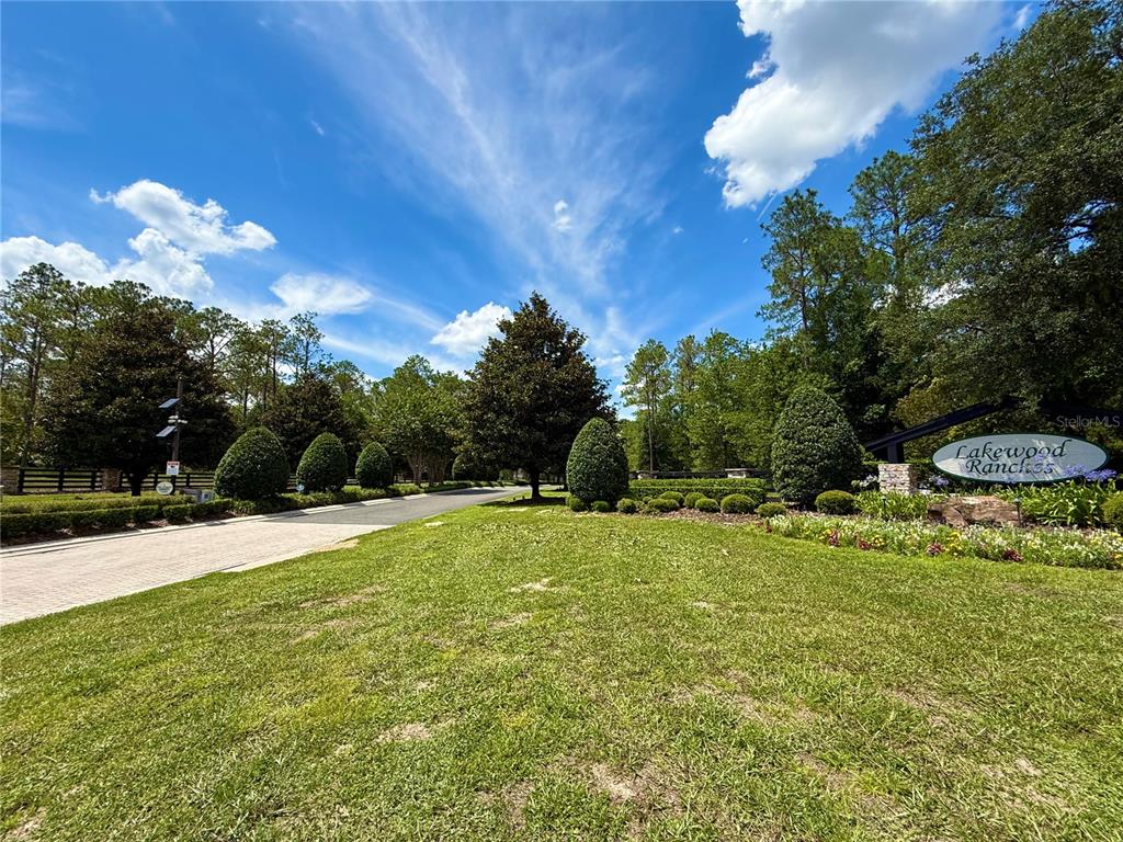 A paved driveway leads toward a landscaped entrance sign for Lakewood Ranches under a blue sky — 35135 PINEGATE TRAIL, EUSTIS, FL