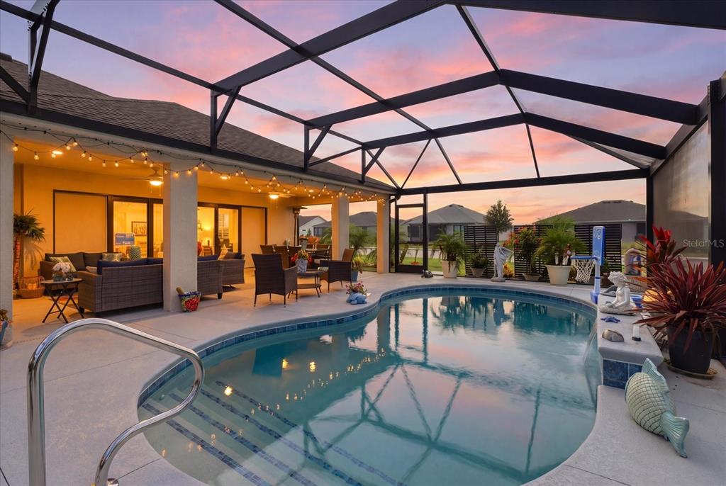 A covered patio features a swimming pool, outdoor seating, and string lights at sunset — 7634 FRANK DIPIETRO DR DRIVE, THE VILLAGES, FL