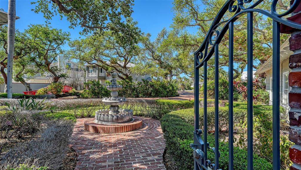 A stone fountain sits on a brick path in a garden viewed through an iron gate — 1249 S BASIN LANE, SARASOTA, FL