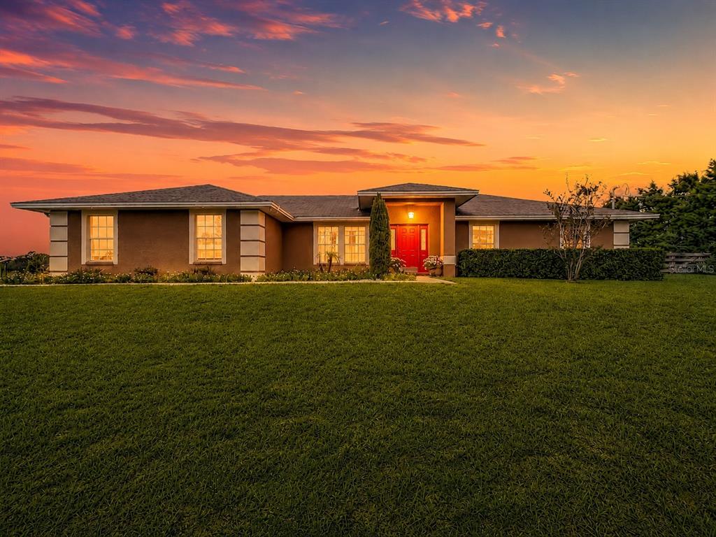 A single-story house with a red front door sits behind a large green lawn at sunset — 18030 BAYAMON AVENUE, POLK CITY, FL