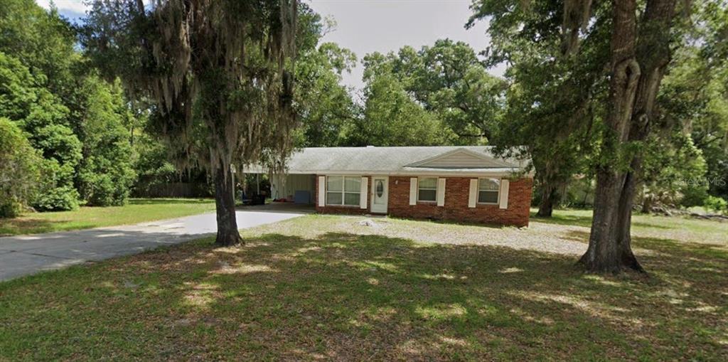 A grassy backyard features mature trees and a white house with a carport — 1965 MANOR WAY, DELAND, FL