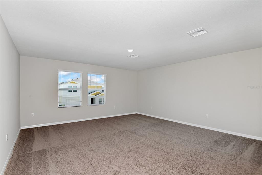 An empty room with beige walls, brown carpeting, and two windows overlooking neighboring houses — 9252 ORCHARD PARK LANE, DADE CITY, FL