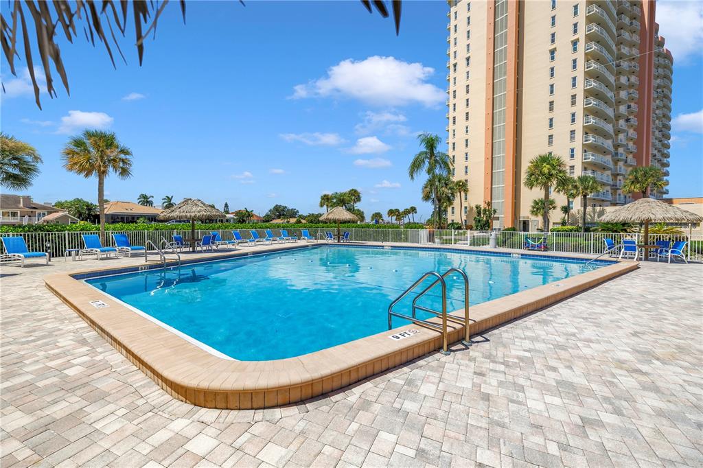 An outdoor swimming pool with lounge chairs and palm trees sits beside a tall building — 4900 BRITTANY DRIVE S UNIT 205, ST PETERSBURG, FL