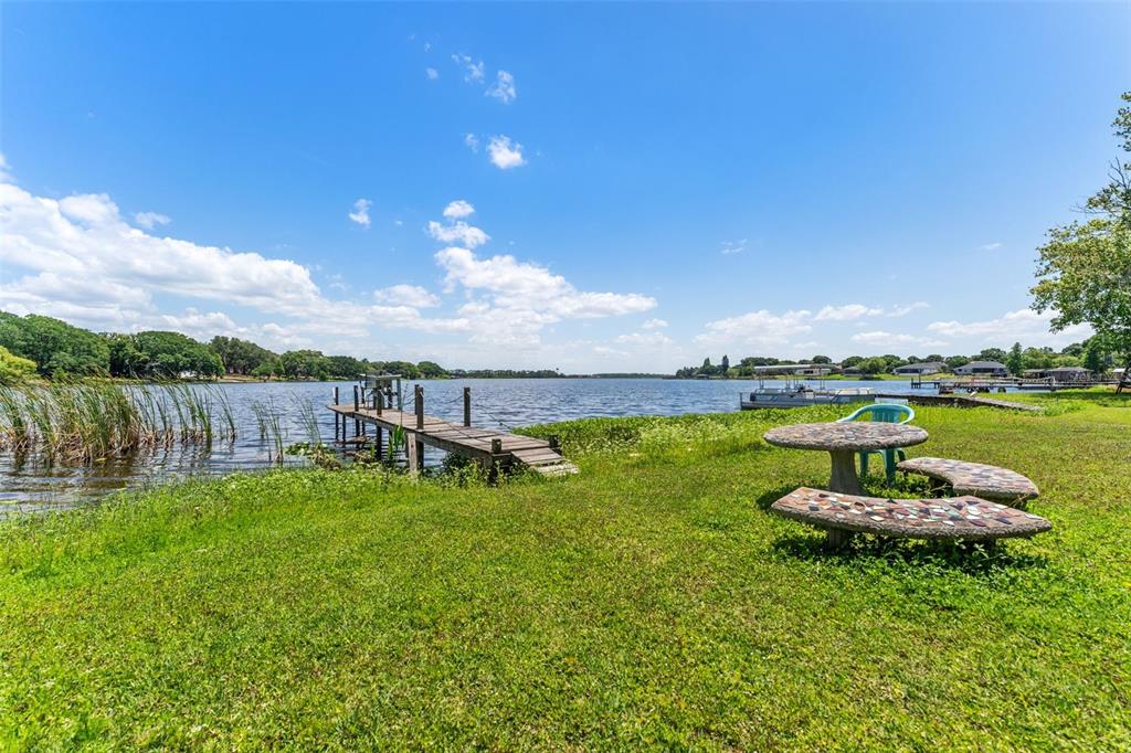 A grassy waterfront yard features a wooden dock, a stone picnic table, and a pontoon boat — 7570 BERKLEY ROAD, POLK CITY, FL