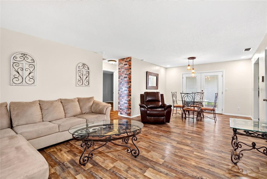 A living and dining area features wood-look flooring, a beige sectional, and a brick accent wall — 27 CEDAR TRACE, OCALA, FL