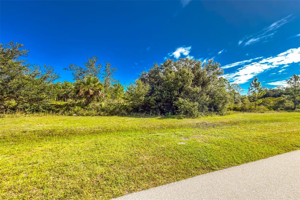 A grassy vacant lot features a paved path in the foreground and dense trees behind — 16550 PRAIRIE CREEK BOULEVARD, PUNTA GORDA, FL