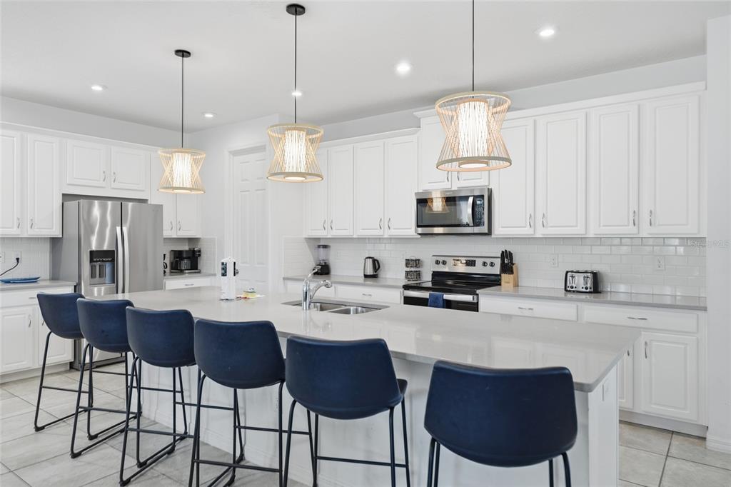 A modern white kitchen features a large island with blue bar stools and pendant lighting — 8967 AMELIA DOWNS TRAIL, KISSIMMEE, FL