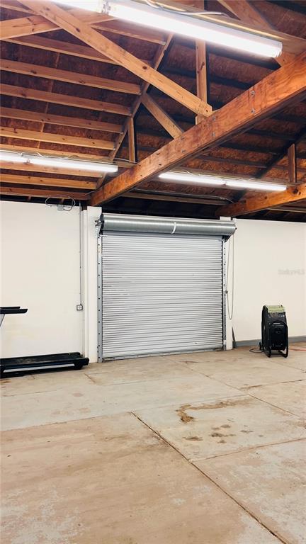 A garage interior features a roll-up metal door, exposed wood rafters, and concrete flooring — 115-119 MICHIGAN AVENUE, DAYTONA BEACH, FL