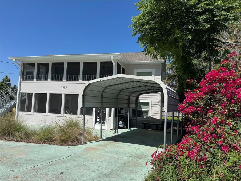 A two-story house with a screened porch and a metal carport sits beside a blooming bougainvillea — 1357 LAKESHORE DRIVE, MOUNT DORA, FL