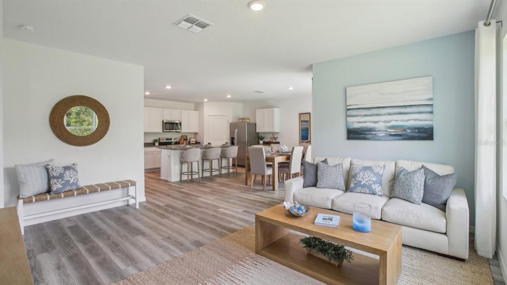 An open-concept living room and kitchen feature light wood flooring and coastal-style decor — 15013 CROSSHALL STREAM DRIVE, WIMAUMA, FL
