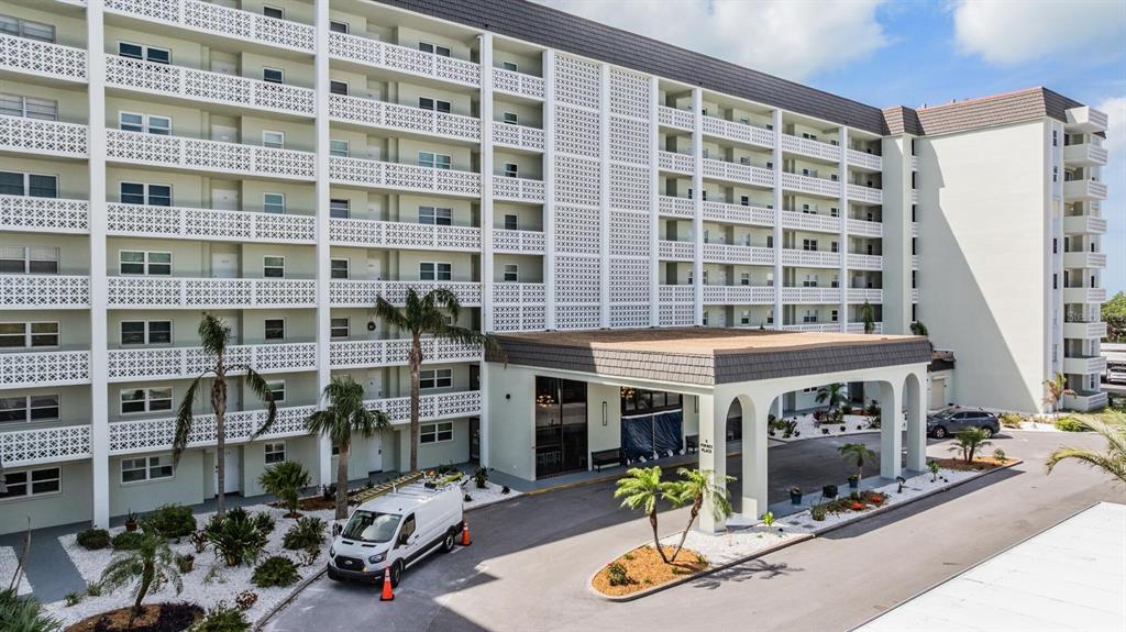 A multi-story apartment building features a covered entrance, balconies, and a paved parking area — 9 FORBES PLACE UNIT 507, DUNEDIN, FL
