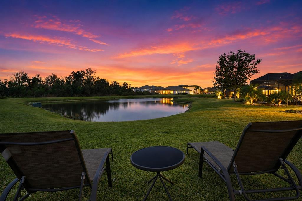 Two lounge chairs and a small table face a pond at sunset near suburban houses — 33564 WILLOW POINT CT, WESLEY CHAPEL, FL