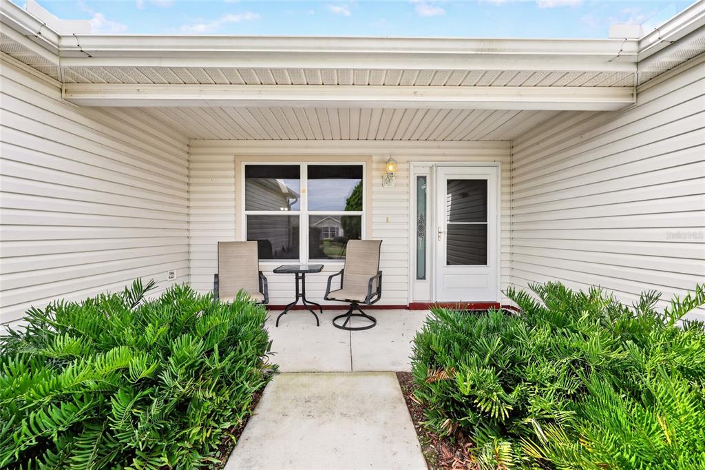 A covered concrete porch features two chairs, a small table, and surrounding green shrubs — 168 PALERMO PLACE, LADY LAKE, FL