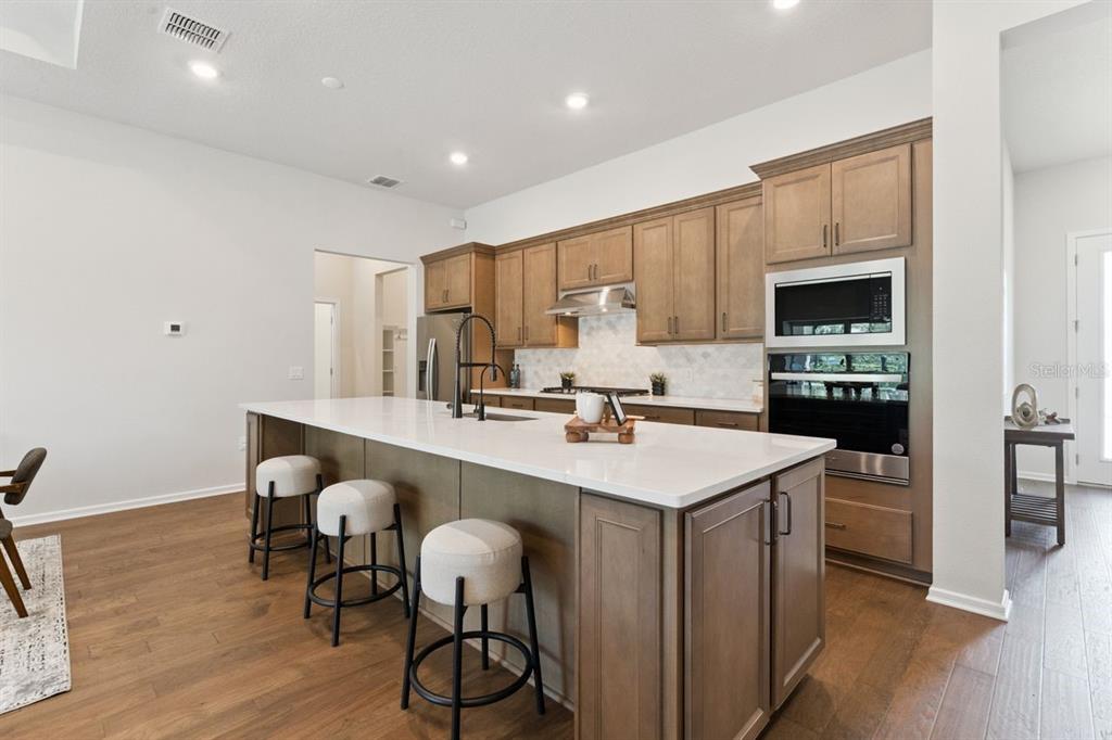A kitchen features a large white quartz island, wood cabinetry, and stainless steel appliances — 15084 CANOPY COVER DRIVE, WINTER GARDEN, FL