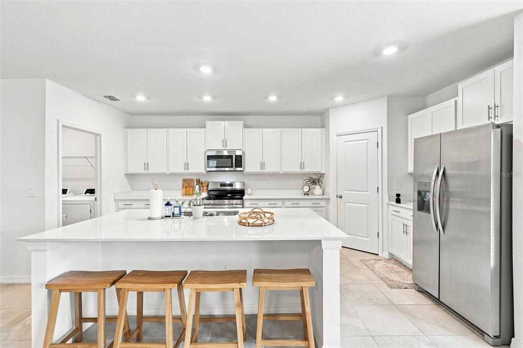 A bright kitchen features white cabinetry, a large central island with wooden stools, and stainless appliances — 13969 LILAC SKY TERRACE, BRADENTON, FL