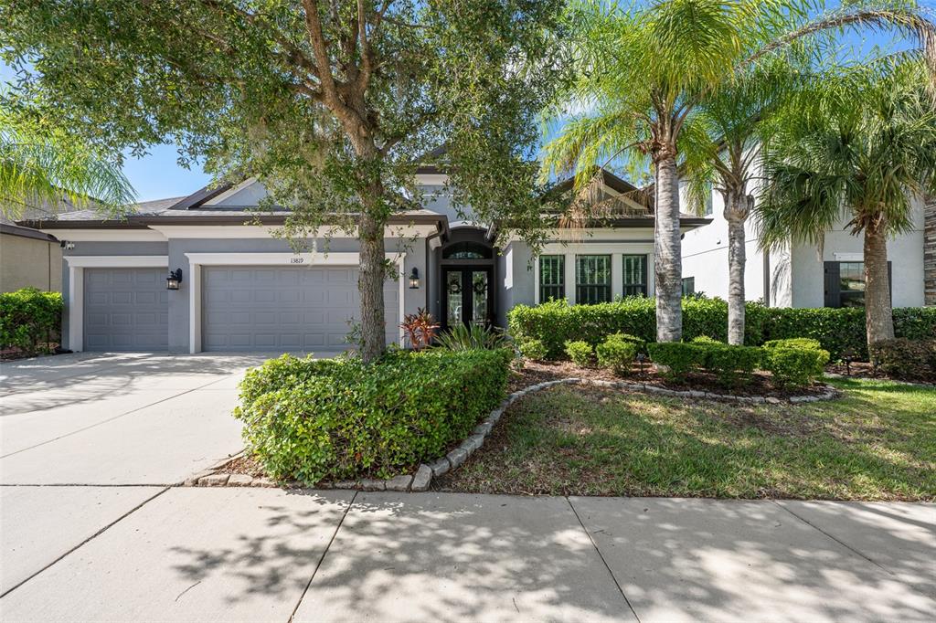 A gray single-story house with a three-car garage, palm trees, and a landscaped front yard — 13819 MOONSTONE CANYON DRIVE, RIVERVIEW, FL