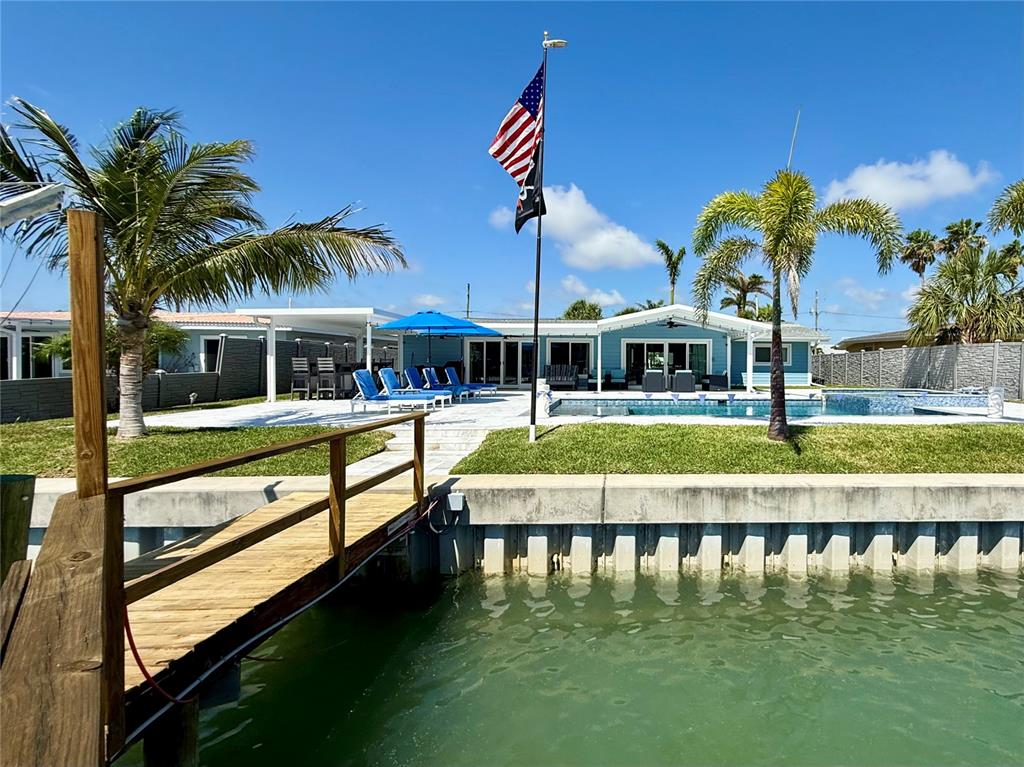 A waterfront home features a swimming pool, patio, and a wooden dock under a blue sky — 12300 4TH STREET E, TREASURE ISLAND, FL