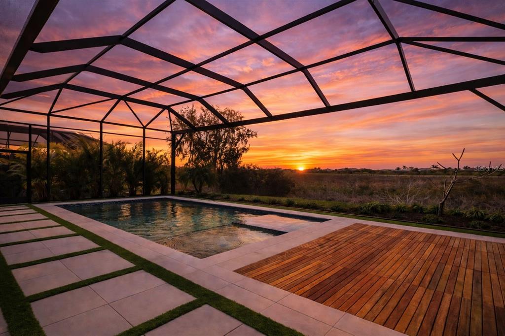A swimming pool with a wooden deck sits inside a screened lanai at sunset — 7982 REDONDA LOOP, BRADENTON, FL