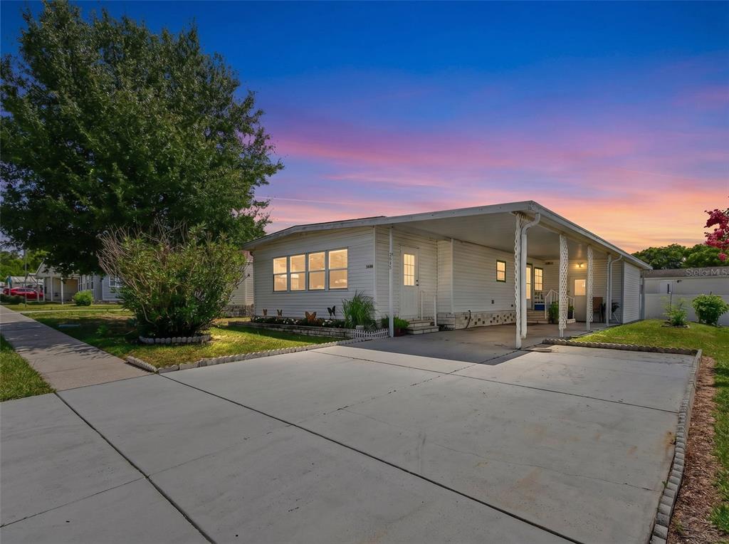 A single-story white mobile home features a concrete driveway and a covered carport at sunset — 3902 JULIE DRIVE, WESLEY CHAPEL, FL