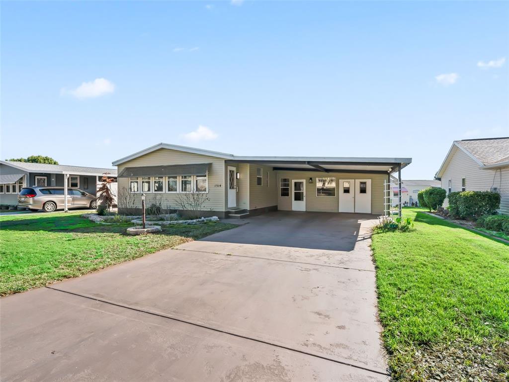 A single-story manufactured home with a concrete driveway and attached carport under a blue sky — 1514 HILLCREST DRIVE, LADY LAKE, FL