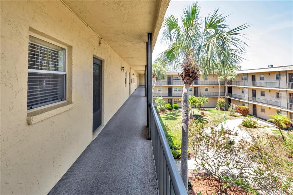 An outdoor apartment walkway overlooks a courtyard with palm trees and a multi-story building — 11485 OAKHURST ROAD UNIT 200-321, LARGO, FL