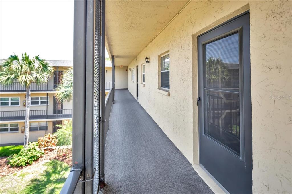 An outdoor apartment walkway features a gray door, textured beige walls, and metal railings — 11485 OAKHURST ROAD UNIT 200-321, LARGO, FL