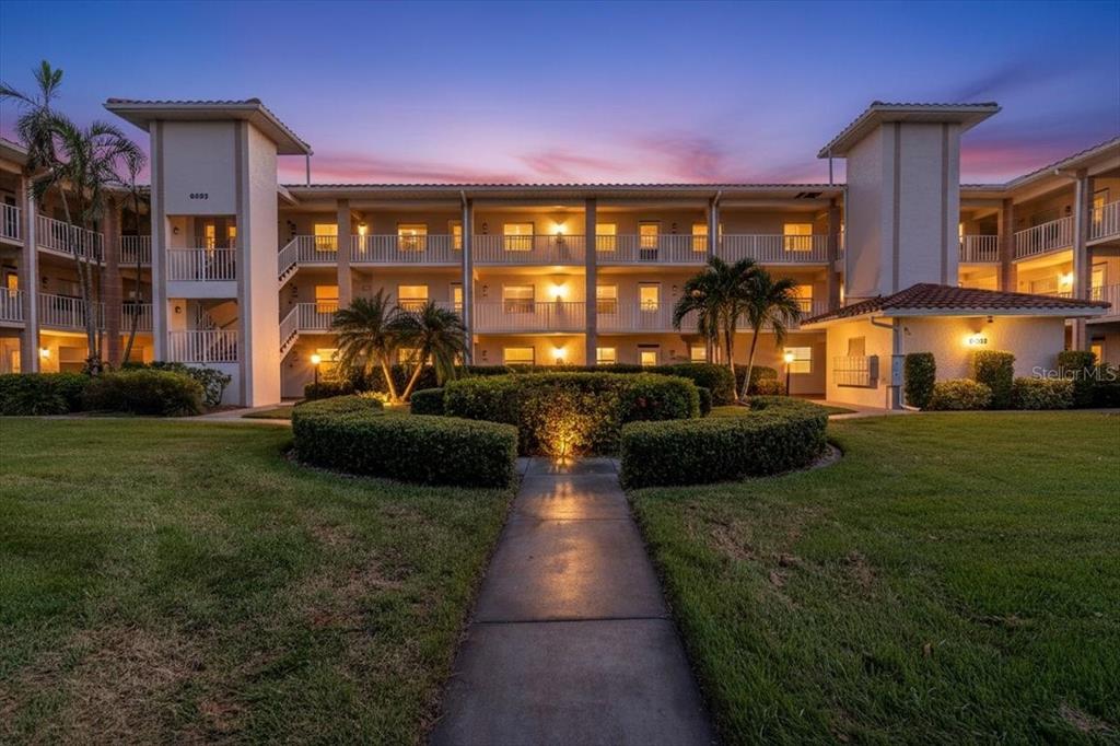 A multi-story apartment building exterior features a central walkway, manicured hedges, and palm trees — 6505 STONE RIVER ROAD UNIT 103, BRADENTON, FL