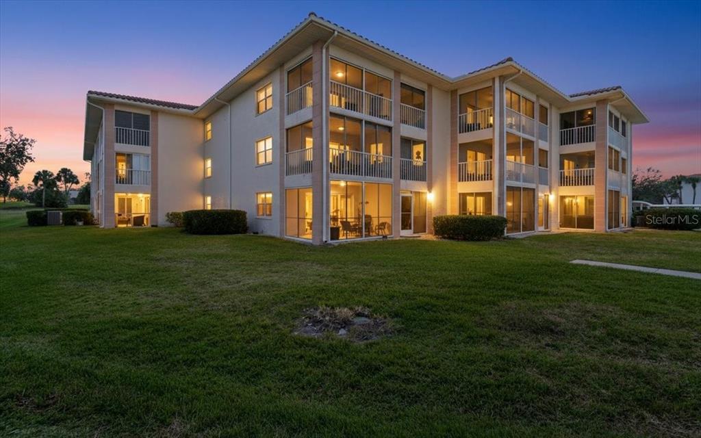A three-story apartment building with screened balconies sits on a grassy lawn at sunset — 6505 STONE RIVER ROAD UNIT 103, BRADENTON, FL