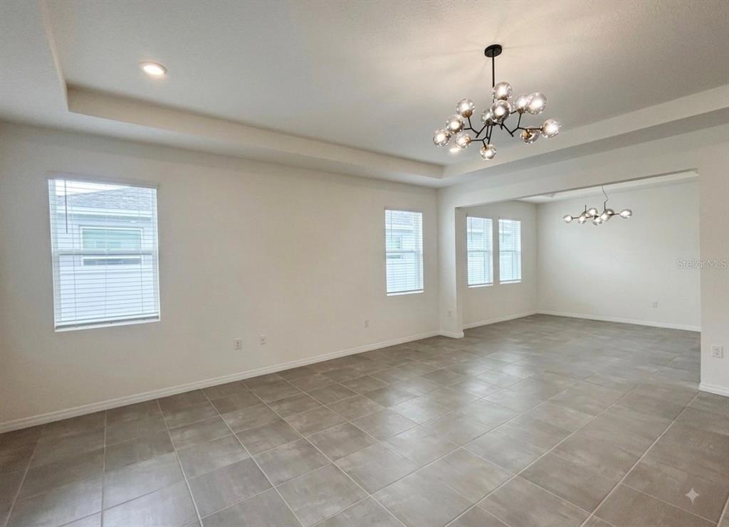 An empty room with gray tile flooring, white walls, tray ceilings, and two modern chandeliers — 5811 SW 80TH LANE, OCALA, FL