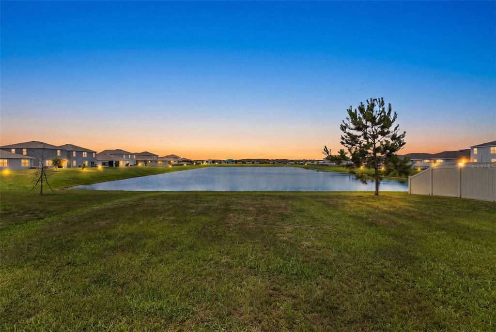 A grassy backyard overlooks a pond at sunset with houses in the distance — 9728 CRESCENT RAY DRIVE, WESLEY CHAPEL, FL