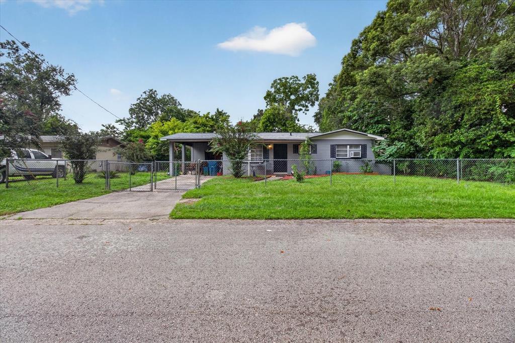 A gray single-story house with a carport and chain-link fence sits behind a paved street — 21385 LEHOUIER DRIVE, BROOKSVILLE, FL