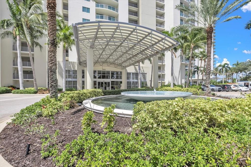 A modern building entrance features a large metal canopy, a circular fountain, and palm trees — 3000 OASIS GRAND BOULEVARD UNIT 2802, FORT MYERS, FL